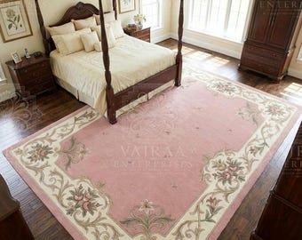 May include: A large rectangular area rug featuring a soft pink center field and a wide cream-colored border. The border is decorated with intricate floral motifs in shades of green, rose, and brown, arranged in symmetrical scrolling patterns.