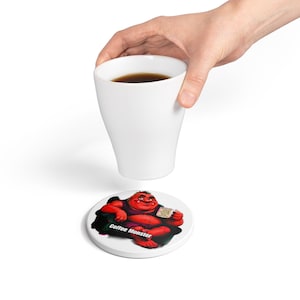 Puede incluir: Una taza de cerámica blanca llena de café oscuro y un posavasos redondo blanco con un personaje de monstruo rojo sentado en una silla negra, sosteniendo una taza pequeña, con el texto Coffee Monster impreso en letras blancas bajo el personaje.