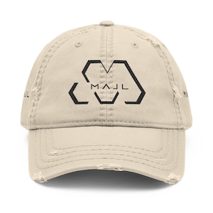 Puede incluir: Una gorra de béisbol beige con aspecto desgastado que presenta un logotipo geométrico negro en el panel frontal con el texto MAJL centrado. La gorra tiene una visera curva con bordes deshilachados y el texto MAJL en los laterales.