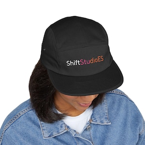 Puede incluir: Una gorra negra de cinco paneles con el texto ShiftStudioES bordado en la parte frontal en letras blancas, rosas y naranjas. La gorra cuenta con una visera plana y una corona estructurada, confeccionada en tela negra lisa con ojales laterales.
