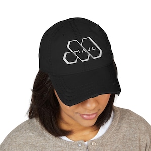Puede incluir: Una gorra de béisbol negra con un acabado desgastado en la visera y la corona. La parte frontal presenta un diseño gráfico blanco que consiste en tres formas hexagonales interconectadas con el texto MAJL en una fuente sans-serif limpia.