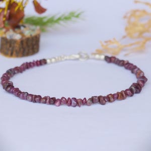 Puede incluir: Una pulsera compuesta por pequeñas cuentas de rubí de color rosa intenso, irregulares y con forma de astilla. La pieza presenta un cierre metálico de tono plateado en el centro, que une los dos extremos del hilo de piedras preciosas en círculo.