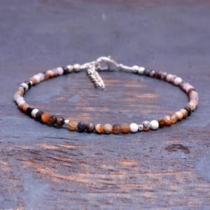 May include: A delicate bracelet composed of small, faceted, multi-colored stone beads in shades of brown, tan, orange, and white. The bracelet features a silver-toned metal lobster claw clasp and an adjustable chain link extension at the back.