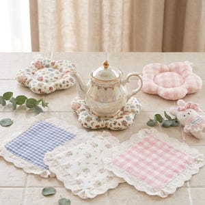 Puede incluir: Una tetera de cerámica con borde dorado sobre un posavasos de tela floral. Alrededor hay dos posavasos de tela con forma de flor, tres posavasos cuadrados con bordes de encaje en cuadros azules, flores y cuadros rosas, y un pequeño conejo de peluche.