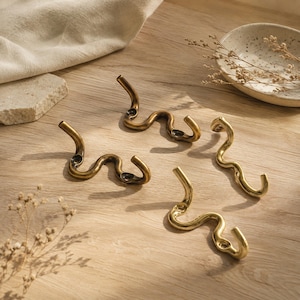 Pode incluir: Quatro ganchos de parede com um design ondulado e sinuoso. Dois ganchos apresentam um acabamento em latão antigo escuro, enquanto os outros dois têm um acabamento dourado brilhante e polido. Cada gancho inclui dois orifícios de montagem.