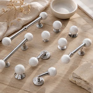 Op de afbeelding: Een collectie meubelbeslag met bolvormige knoppen en handgrepen van wit marmer op gepolijste zilverkleurige metalen voetjes. De set bevat diverse lengtes ladehandgrepen en individuele ronde knoppen, allemaal met een consistente witte steenafwerking.