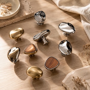 May include: A collection of eight cabinet knobs in various finishes, including polished chrome, brushed gold, and wood-inlaid metal. Each knob features an irregular, organic, pebble-like shape with a central mounting post for attachment to furniture.