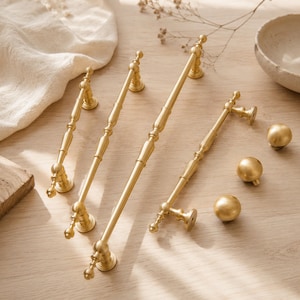 May include: A collection of brushed gold cabinet hardware including three elongated bar pulls with decorative spindle detailing and three matching spherical knobs. The metal hardware features a smooth, matte finish and rounded mounting bases.