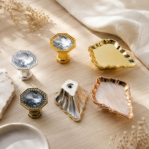 May include: A collection of cabinet hardware including three octagonal knobs with faceted crystal centers in silver, gold, and bronze tones, alongside two shell-shaped pulls in solid gold and white with a gold rim, and one silver fluted knob.
