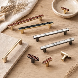 May include: A collection of cabinet hardware including long bar pulls and T-shaped knobs. The pieces feature gold, black, and dark metal finishes with inlaid details in wood grain, solid black, and white marble patterns, arranged on a wooden surface.