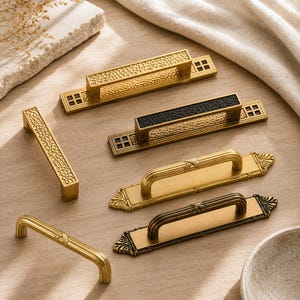 Puede incluir: Una colección de seis tiradores de armario de metal con varios acabados. Dos presentan texturas doradas martilladas con placas finales cuadradas, una con incrustación negra. Dos tienen asas arqueadas doradas lisas sobre placas rectangulares.
