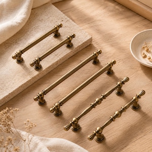 Könnte beinhalten: Eine Sammlung von sechs Schrankgriffen aus antikem Messing mit metallischem Finish. Vier Griffe haben ein glattes, zylindrisches Stangendesign mit abgerundeten Enden, während zwei Griffe eine dekorative, mehrfach segmentierte Perlenstruktur aufweisen.