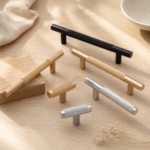 May include: A collection of five cabinet pulls and knobs in various finishes including matte black, brushed gold, and silver. Each piece features a cylindrical bar design with a central textured knurled grip section and two mounting posts.