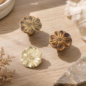 May include: Three metal cabinet knobs shaped like flowers with scalloped edges. Two knobs feature a dark, antique brass finish with visible patina, while the third knob has a bright, polished gold finish. Each knob has a central circular detail.