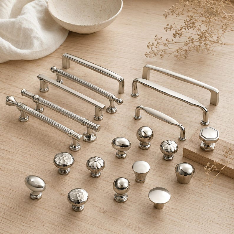 May include: A collection of silver-toned metal cabinet hardware including various drawer pulls and knobs. The set features smooth, hammered, and fluted textures in round, oval, and bar shapes, all designed for furniture or cabinetry installation.