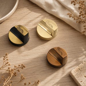 May include: Three circular cabinet knobs with a raised T-shaped handle. One knob is matte black with a gold accent, one is solid brushed gold, and one is solid brushed bronze. Each handle features a small engraved fan-like pattern on the top.