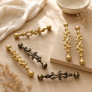 May include: A collection of five metal cabinet pulls featuring a vine and leaf design. Three pulls have a bright gold finish, while two pulls have a dark, antiqued bronze finish. Each pull has a central twisted vine motif with small leaves.