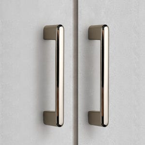 May include: Two vertical cabinet pulls with a polished metallic finish and a smooth, rounded rectangular profile. Each handle is mounted onto a light gray, textured surface, featuring a clean, straight design with rounded ends and a reflective surface.