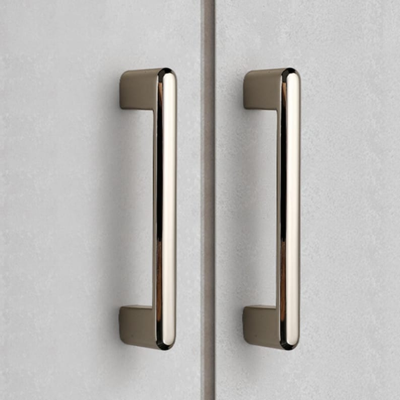 May include: Two vertical cabinet pulls with a polished metallic finish and a smooth, rounded rectangular profile. Each handle is mounted onto a light gray, textured surface, featuring a clean, straight design with rounded ends and a reflective surface.