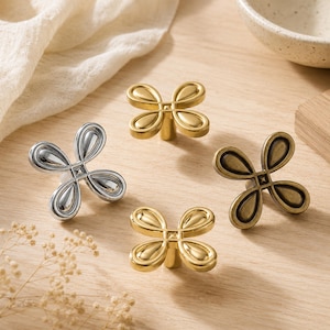 May include: Four metal cabinet knobs shaped like four-petaled flowers. The set includes one polished silver knob, one brushed gold knob, one bright gold knob, and one antique bronze knob, each featuring a central square detail and rounded petals.