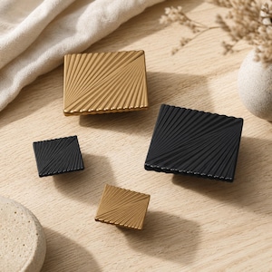 May include: Four square cabinet knobs in two sizes and two colors. Two knobs are matte gold and two are matte black. Each knob features a textured surface with a radiating fan-like pattern of diagonal lines extending from one corner.