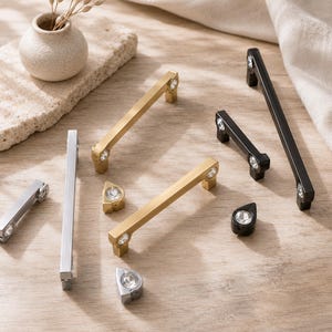 Op de afbeelding: Een collectie meubelbeslag bestaande uit lange stanggrepen en kleine knoppen in goud-, zilver- en zwarte afwerkingen. Elk stuk is voorzien van heldere, gefacetteerde kristallen accenten aan de uiteinden of in het midden van het metaal.