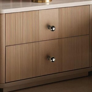 May include: A light wood nightstand featuring two drawers with vertical fluted paneling and smooth center sections. Each drawer is equipped with a single polished silver spherical knob. The unit has a flat top surface and a solid base frame.