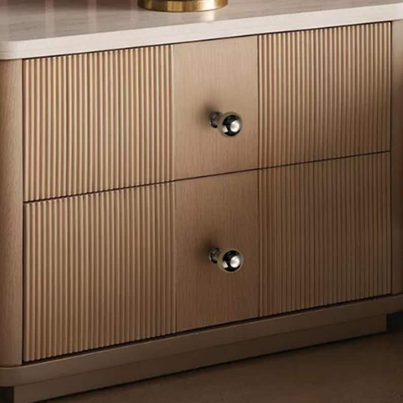 May include: A light wood nightstand featuring two drawers with vertical fluted paneling and smooth center sections. Each drawer is equipped with a single polished silver spherical knob. The unit has a flat top surface and a solid base frame.