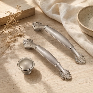 May include: Two brushed silver metal drawer pulls with ornate, scroll-like detailing at each end and one matching round brushed silver cabinet knob. The hardware features a smooth, matte finish and a curved design for easy gripping and installation.