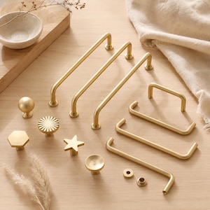 May include: A collection of brushed gold cabinet hardware including three long arched pulls, three shorter arched pulls, and several decorative knobs in round, hexagonal, star, and fluted shapes, along with two small circular metal washers.