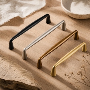 Pode incluir: Quatro puxadores de armário em metal dispostos em fila, com acabamentos distintos em preto mate, prata escovada, bronze antigo e ouro acetinado. Cada puxador apresenta um design de barra arqueada lisa com suportes cónicos nas extremidades.