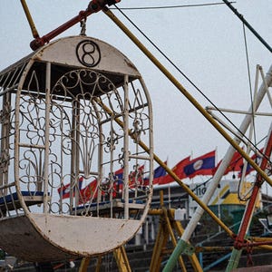 May include: A white, oval-shaped Ferris wheel passenger car featuring an intricate metal scrollwork design and the number eight enclosed in a circle at the top. The structure is suspended from a yellow and red metal frame against a cloudy sky.