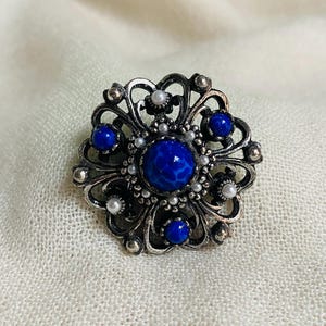 May include: A circular silver-toned brooch featuring an intricate openwork metal frame. The centre is set with a large, round, mottled blue stone, surrounded by smaller blue cabochons and tiny white faux pearls arranged in a floral pattern.