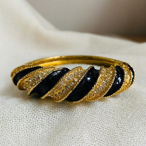 May include: A hinged bangle bracelet featuring a twisted design with alternating bands of glossy black enamel and gold-tone metal. The gold-tone sections are textured with a rope-like pattern and are embedded with small, clear, round rhinestones.