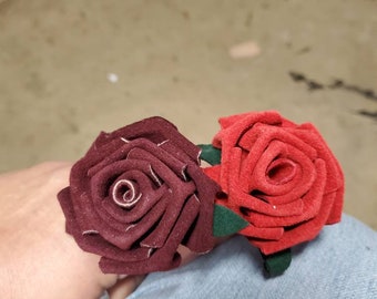 Op de afbeelding: Twee haaraccessoires met rozen van stof. Eén is diep bordeauxrood van kleur, de andere is felrood. Beide hebben groene bladeren aan de basis. De rozen zijn gemaakt van een zacht materiaal en zijn waarschijnlijk handgemaakt.