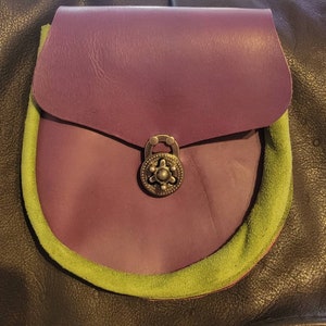 Op de afbeelding: Een paarse leren tas met een groene su&egrave;de rand en een messing sluiting. De tas heeft een klep met een decoratief metalen ontwerp.