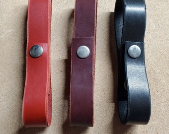 Op de afbeelding: Drie leren sleutelhangers in rood, bordeaux en zwart. Elke sleutelhanger is een lus met een metalen drukknoopsluiting. De sleutelhangers worden gepresenteerd op een lichtbruin oppervlak.