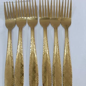 Può includere: Un set di sei forchette in metallo color oro disposte in fila. Ogni forchetta presenta una testa a quattro rebbi e un manico decorato con un motivo floreale in rilievo che si estende dal collo fino alla base arrotondata del manico.
