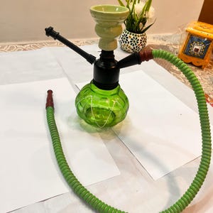 May include: A hookah featuring a translucent green glass base with a ribbed texture, a black central stem, and a light green ceramic bowl. A long, flexible green hose with a brown mouthpiece is attached to the side of the black stem assembly.