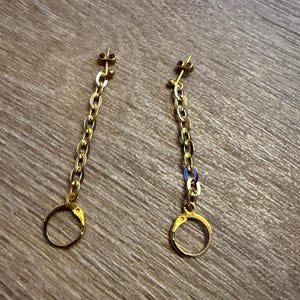Pode incluir: Um par de brincos pendentes em tom dourado com um design delicado de elos de corrente. Cada brinco consiste num pequeno pino circular no topo, ligado a uma corrente vertical de elos ovais, terminando num pequeno pendente circular.