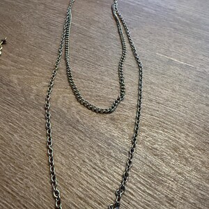 Könnte beinhalten: Eine lange silberfarbene Metallkettenhalskette mit zweilagigem Design. Die Kette besteht aus kleinen, gleichmäßigen ovalen Gliedern und endet unten mit zwei ineinandergreifenden kreisförmigen Ringen, die als dekorativer Blickfang dienen.