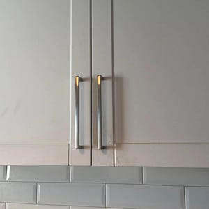 May include: Two white kitchen cabinet doors featuring a shaker-style frame design. Each door is fitted with a long, vertical, brushed silver metal handle. The cabinets are positioned above a backsplash of light grey rectangular subway tiles.
