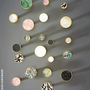 May include: A collection of round cabinet knobs with brass rims, featuring various inlaid patterns including solid white, black glitter, iridescent abalone shell, marbled pink, and green swirls. The text BRASS HARDWARE is visible on the left side.