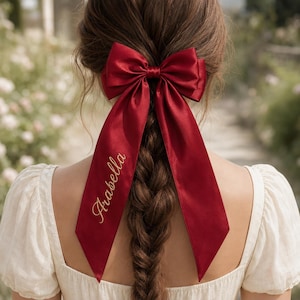 Può includere: Un fiocco per capelli in raso rosso scuro con lunghi nastri pendenti. Sul nastro sinistro è ricamato il nome Arabella in filo dorato. Il fiocco è fissato al centro e poggia su capelli castani intrecciati, indossato con un top bianco.