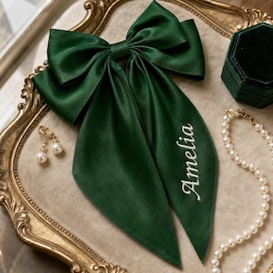 Peut inclure: Un grand nœud pour cheveux en satin vert foncé avec de longs pans tombants. Le prénom Amelia est brodé en lettres cursives blanches sur le pan droit du nœud. Le tissu présente une finition lisse et réfléchissante avec des plis marqués.