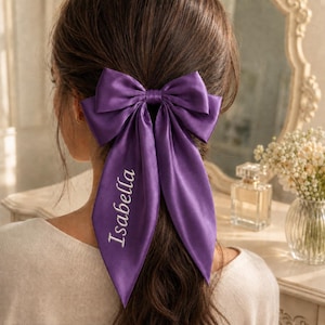 May include: A deep purple satin hair bow featuring two long, pointed tails. The name Isabella is embroidered in white script lettering on the left tail of the bow. The accessory is secured in dark brown hair, showcasing a smooth, reflective fabric finish.