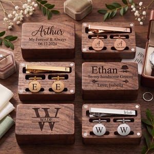 Könnte beinhalten: Drei rechteckige Holzschatullen mit personalisierten Krawattenklammern und Manschettenknöpfen. Jedes Set ist mit individuellen Gravuren versehen, darunter Namen wie Arthur, Ethan und Wade sowie Daten und Initialen. Die Accessoires sind in Roségold, Gold und Silber gehalten.