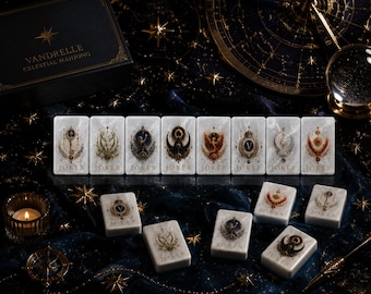 Könnte beinhalten: Ein Set aus weißen, rechteckigen Mahjong-Spielsteinen mit marmorierter Textur, die jeweils mit komplexen himmlischen Illustrationen und dem Wort JOKER am unteren Rand versehen sind. Die Steine liegen neben einer schwarzen Box mit der Aufschrift VANDRELLE CELESTIAL MAHJONG.