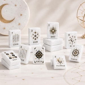 Puede incluir: Un conjunto de fichas rectangulares blancas con ilustraciones y texto en dorado y negro con tem&aacute;tica celestial. Las fichas presentan varios s&iacute;mbolos, incluyendo br&uacute;julas, lunas y estrellas, con etiquetas como DOT, BAM, CRAK, JOKER, FLOWER y NORTH.