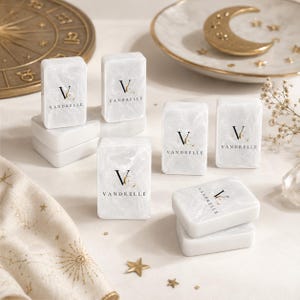 Puede incluir: Una colecci&oacute;n de bloques rectangulares con efecto de m&aacute;rmol blanco que presentan el texto negro VANDRELLE y un logotipo. Los bloques est&aacute;n dispuestos en pilas e individualmente sobre una superficie, acompa&ntilde;ados de una rueda zodiacal dorada y un plato de media luna.